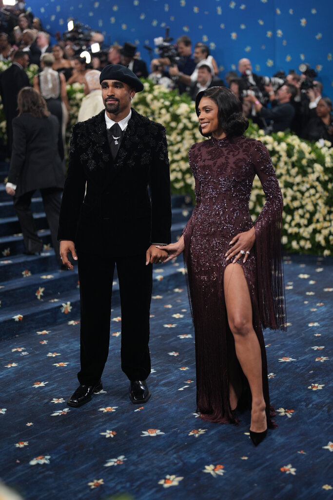 Jalen Hurts, wearing a black suit with embellishments along the shoulders and a black beret, poses with Bry Burrows, wearing a maroon dress with beading, fringe detailing along the arms and a high-cut slit.