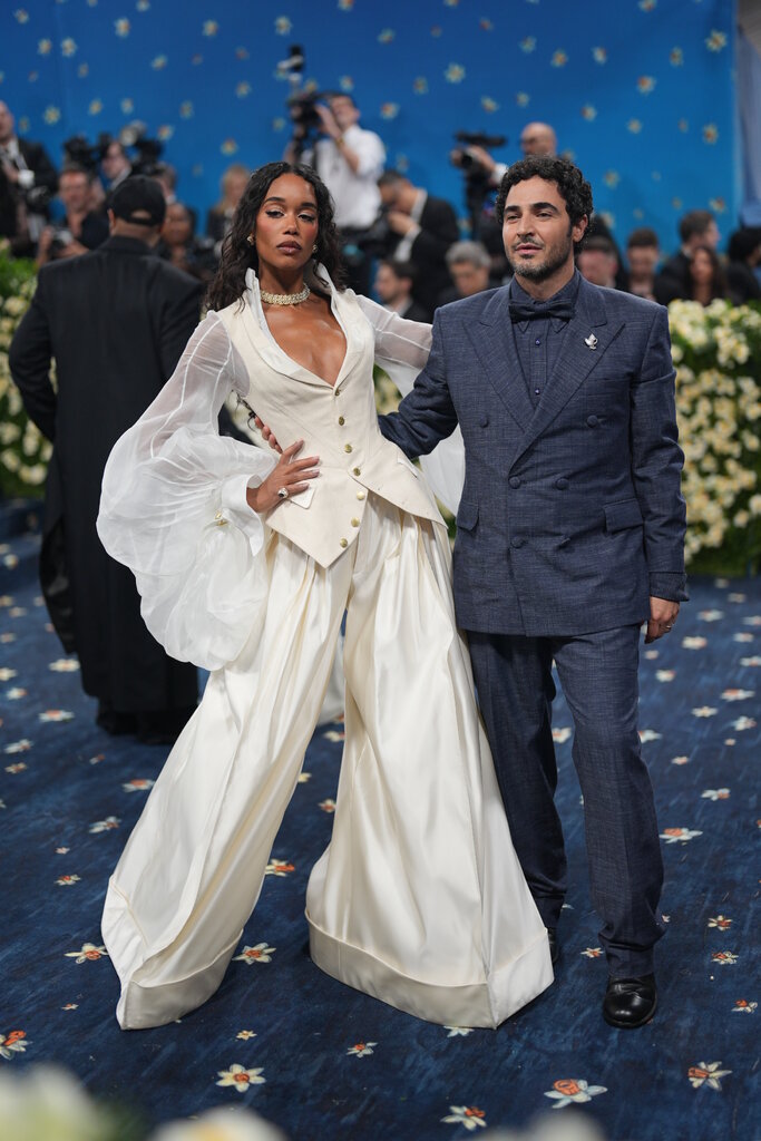Laura Harrier, wearing a low-cut cream blouse with exaggerated sleeves overtop extra-wide wide-leg trousers, poses with Zac Posen, wearing a blue double-breasted suit.