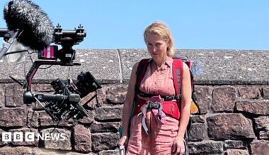 The actor Gillian Anderson looks towards the photographer during the filming of The Salt path in Minehead. She is wearing a pink vest and shorts and has a red rucksack strapped to her back and waist. She has blond hair pulled back in a ponytail and is standing in front of a high grey brick wall with a camera on a tripod pointed at her.