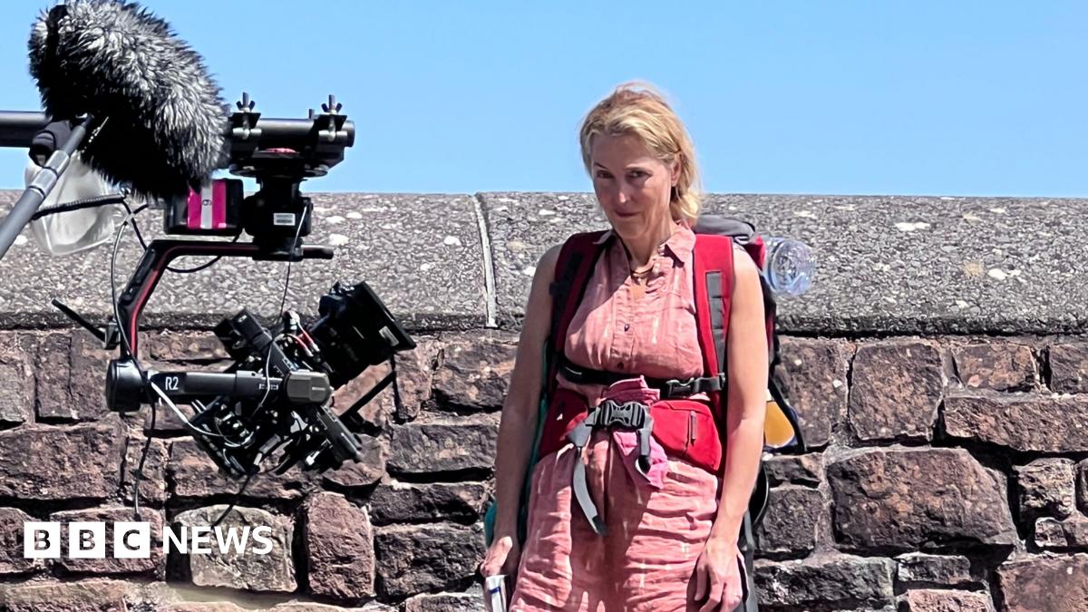 The actor Gillian Anderson looks towards the photographer during the filming of The Salt path in Minehead. She is wearing a pink vest and shorts and has a red rucksack strapped to her back and waist. She has blond hair pulled back in a ponytail and is standing in front of a high grey brick wall with a camera on a tripod pointed at her.