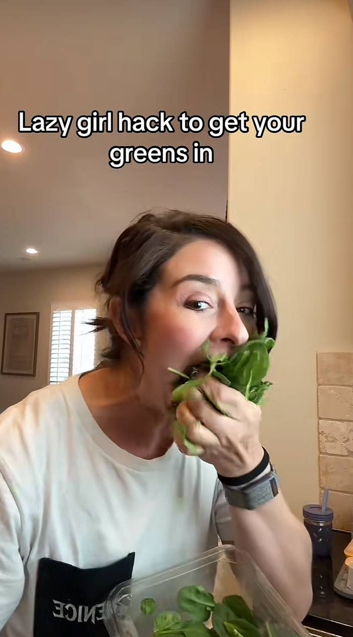 Person eating a handful of fresh greens from a container in a kitchen, with text "Lazy girl hack to get your greens in" above