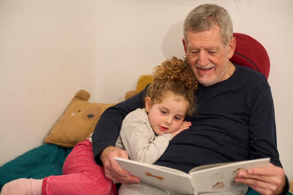 Older man reading a book to a young child, both sitting comfortably. Child leans against the man, listening intently. Cozy atmosphere
