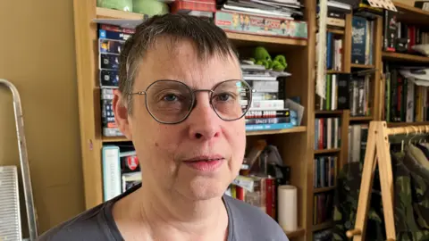 BBC Susan, a woman, with grey short hair, wearing glasses stands in front of shelving filled with books