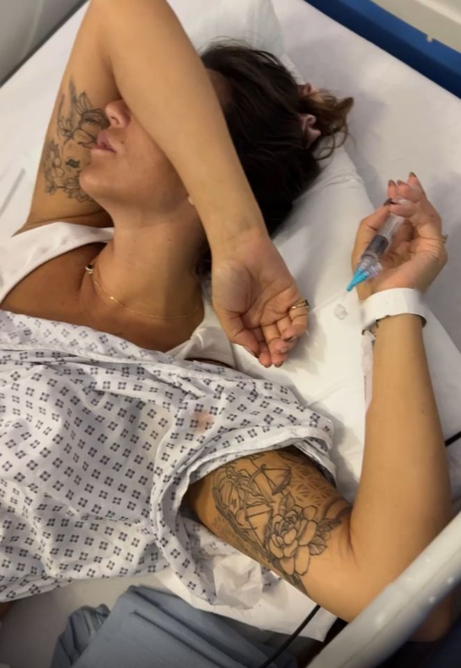 A woman in a hospital gown lies in a hospital bed, holding a syringe.