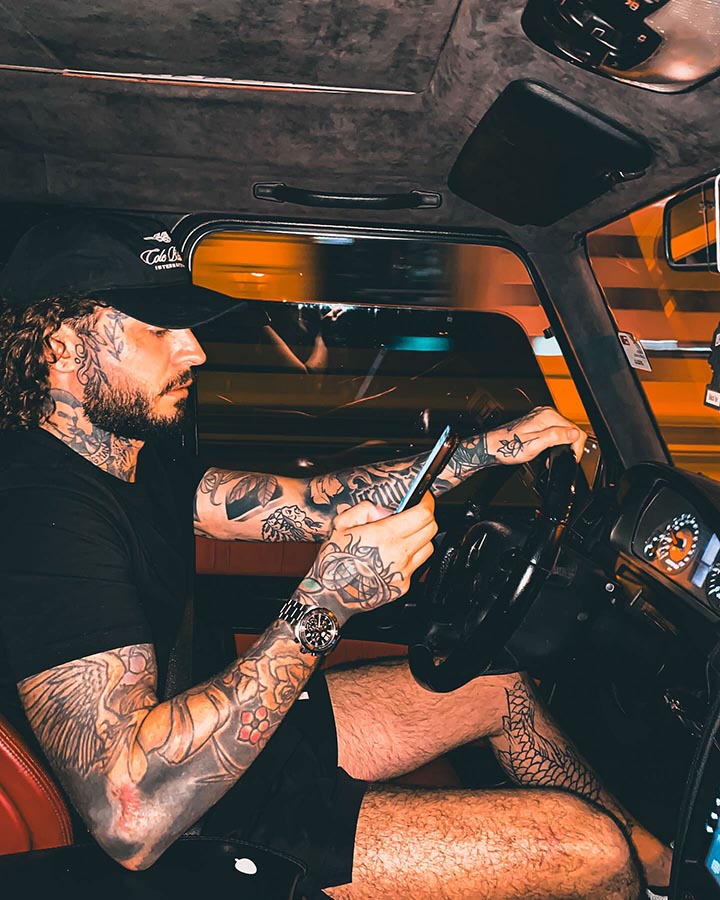 A tattooed man driving a car and using a phone.