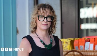 Laura has shoulder length curly blonde hair with a curly fringe. She is wearing black framed glasses and a black vest top. She has a large green necklace on.