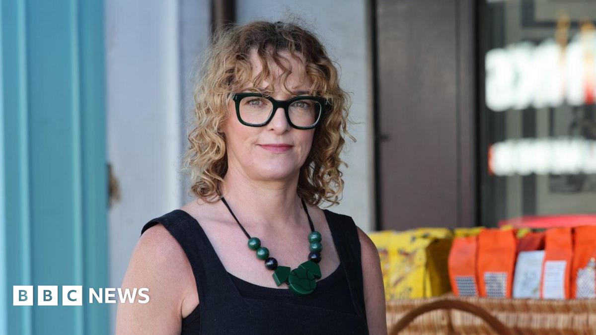Laura has shoulder length curly blonde hair with a curly fringe. She is wearing black framed glasses and a black vest top. She has a large green necklace on.