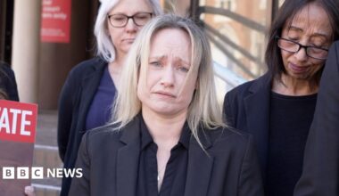 A woman with blonde hair wearing all black. She looks devastated and is holding back tears. There are people standing behind her also looking very upset.