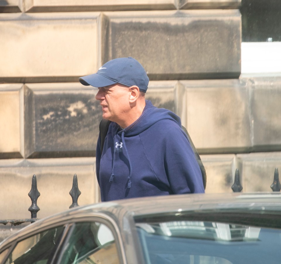 Man in a blue hoodie and baseball cap walking near a car.
