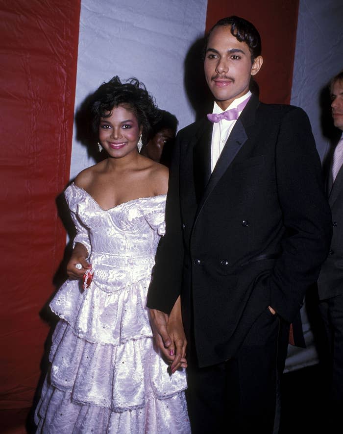 A woman in an off-shoulder, ruffled gown and a man in a tuxedo with a bow tie are holding hands at a formal event
