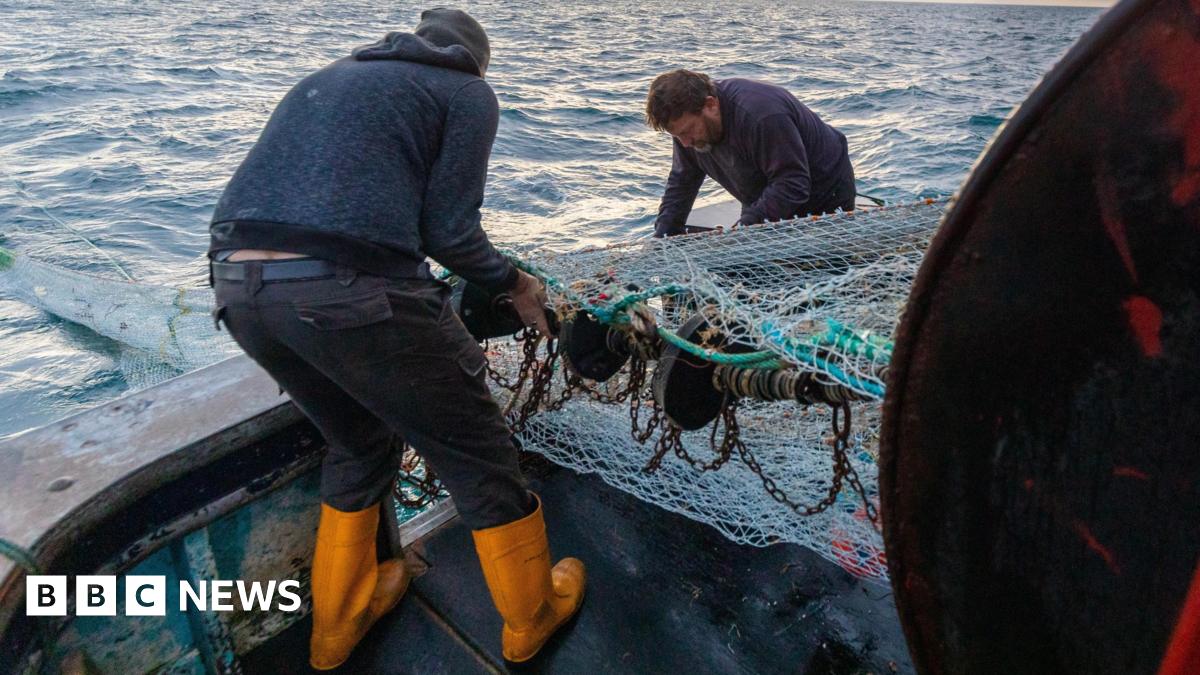 Two fishermen on a boat putting out a fishing net from their boat for their first catch of the day in the English Channel, off the south coast of East Sussex, with waves in the background and soft light from the rising sun.
