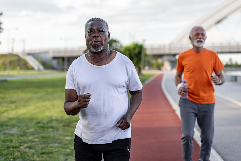 Two men jogging on a track, one in a white shirt and the other in an orange shirt, outdoors on a sunny day