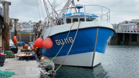 BBC A photo of a fishing boat that is white and blue. It is positioned at a harbour on a grey, cloudy day. 