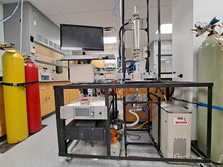 Photo of a chemistry lab space with colorful gas bottles. At the center of the image is a microwave reactor connected by a waveguide to a microwave source.