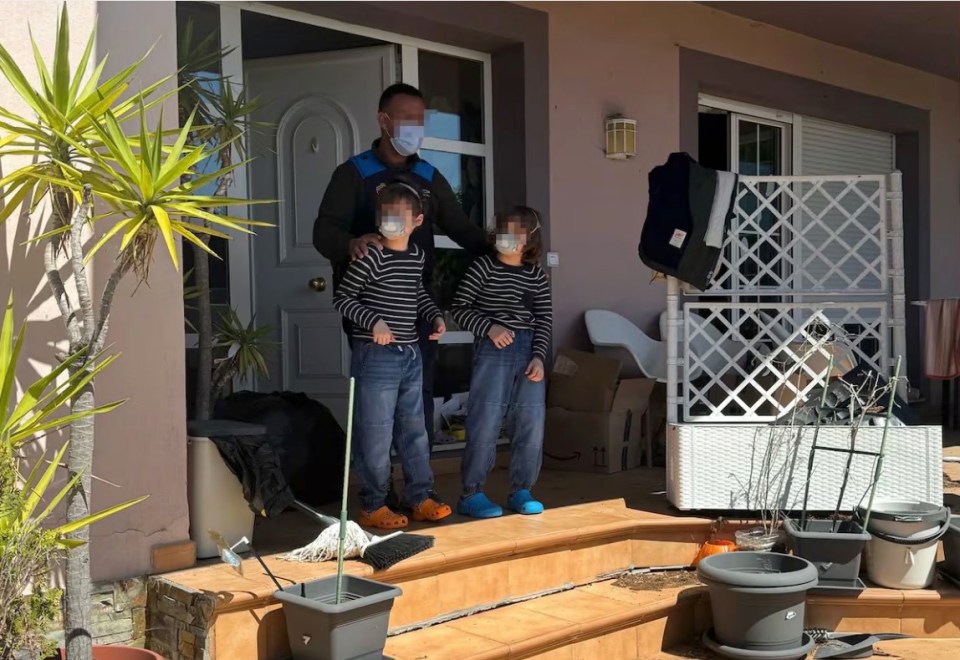 A masked man stands with two masked children on a porch, surrounded by clutter.