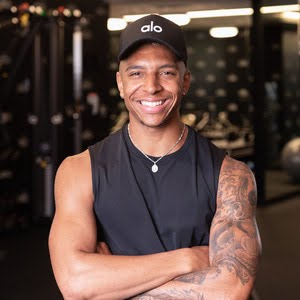 Smiling man with his arms crossed, wearing black tank top, pendent necklace and black cap with 
