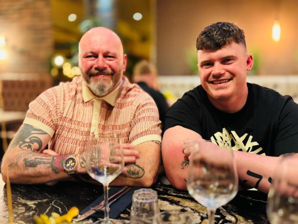 A young man and older man with tattoos sit at a table with wine glasses.