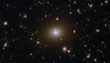 A field full of distant galaxies on a dark background. Most of the galaxies are very small, but there are a few larger galaxies and some stars where detail is visible. In the very center is an elliptical galaxy with a brightly glowing core and a broad disk. A reddish, warped, ring of light, thicker at one side, surrounds its core. A small galaxy intersects the ring as a bright dot.