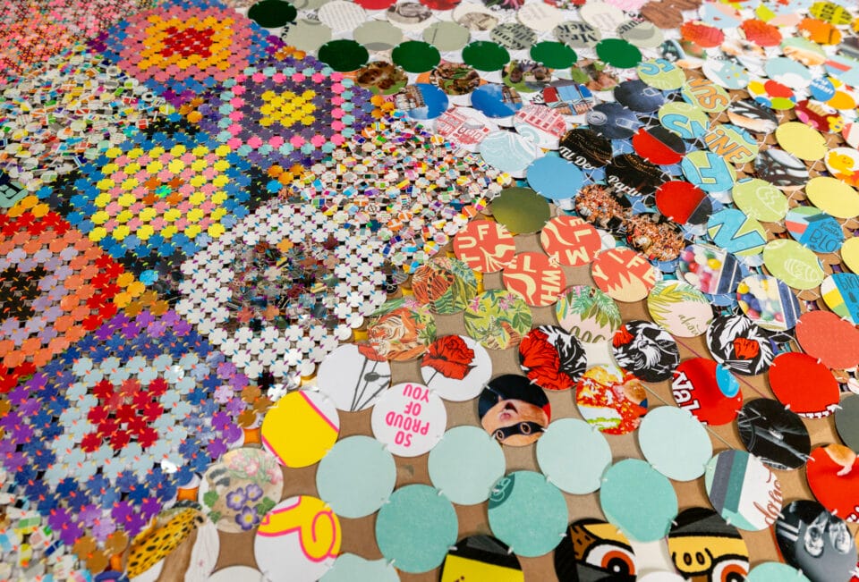 a detail of an artwork comprising cloth made from woven and connected pieces of plastic and paper, creating colorful patterns