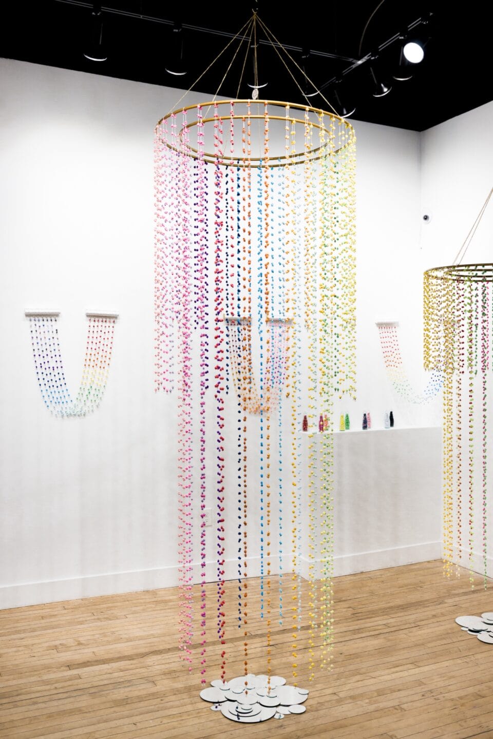 a suspended installation of numerous colorful bead-like pieces complement a number of smaller wall-hung works also made with colorful draping strands of found material