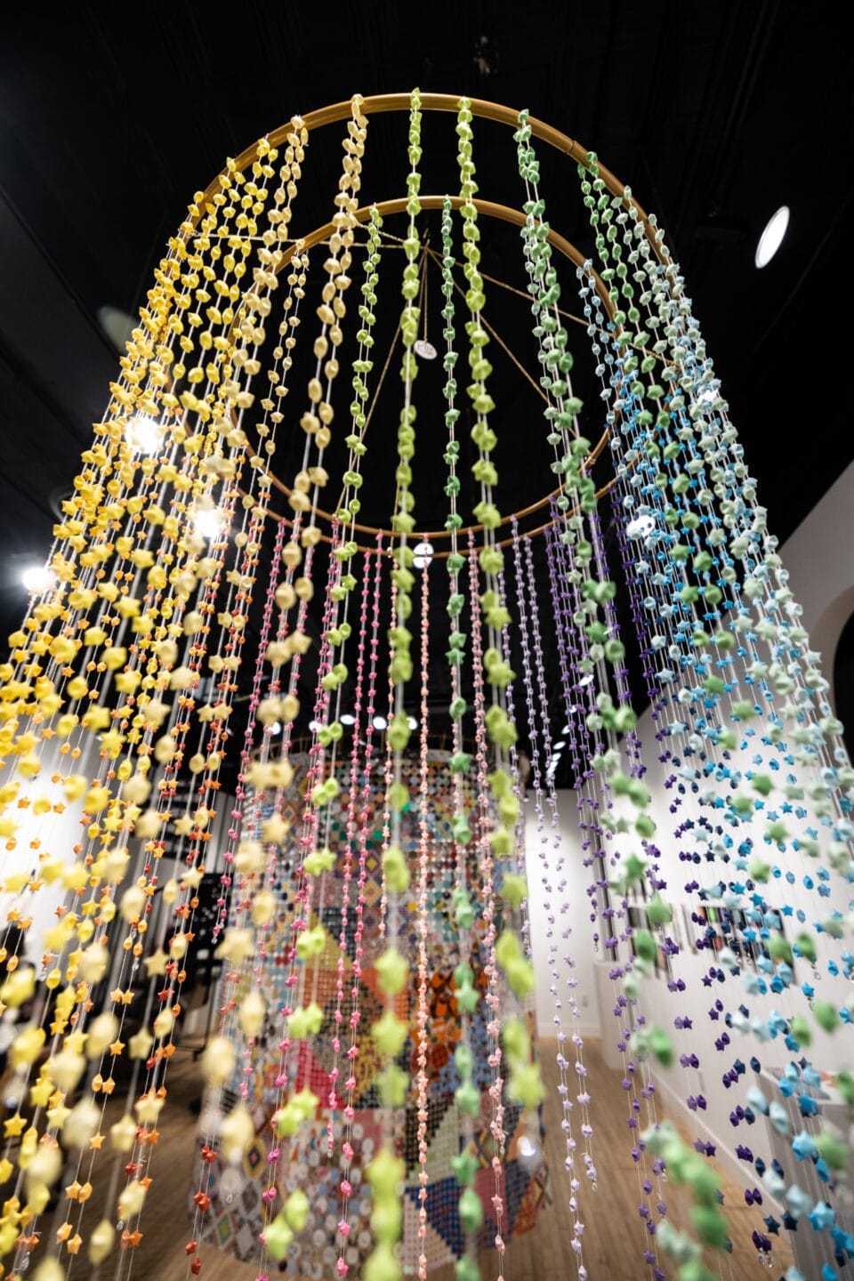 an installation view of a suspended sculpture in a gallery, looking upward, comprising numerous strands of colorful beds