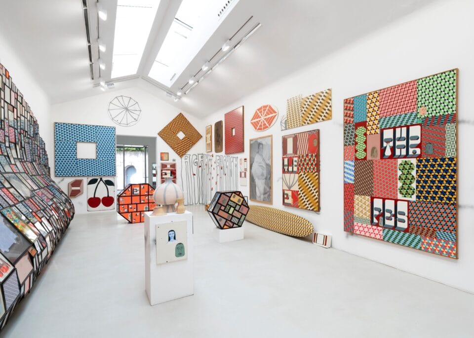 installation view of an exhibition in a white-walled gallery space with numerous prints, paintings, and sculptures in a variety of shapes and colors all over the walls