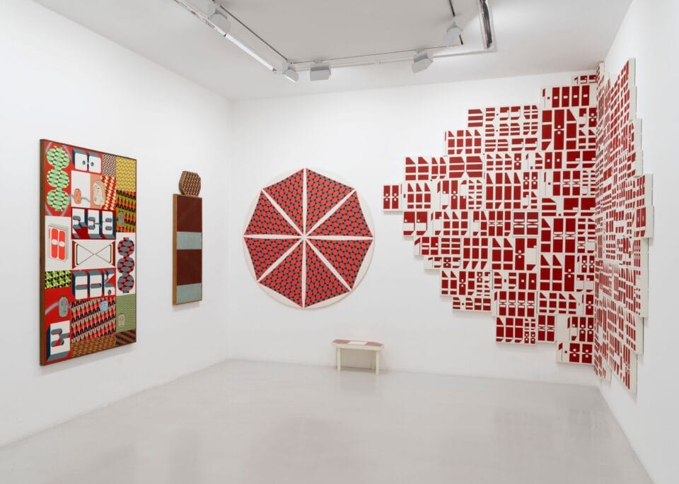 installation view of an exhibition in a white-walled gallery space with numerous prints, paintings, and sculptures in a variety of shapes and colors all over the walls