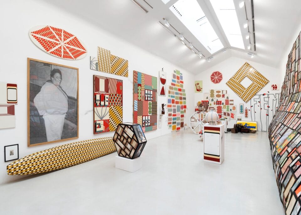 installation view of an exhibition in a white-walled gallery space with numerous prints, paintings, and sculptures in a variety of shapes and colors all over the walls
