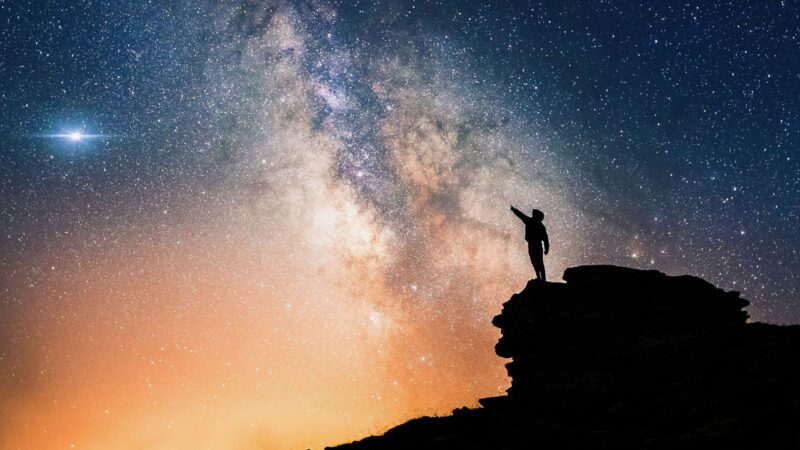 Astronomers respond: lone person standing on top of rock, pointing with hand towards the Milky Way and the vast expanse of universe.