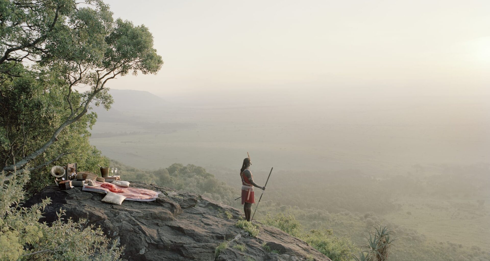 a Masai warrior stands on a hillside, looking out over a hazy landscape, with a pink picnic blanket on the rocks behind him