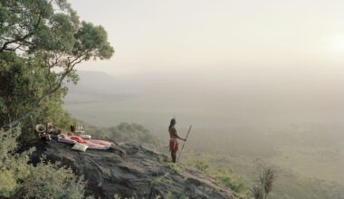 a Masai warrior stands on a hillside, looking out over a hazy landscape, with a pink picnic blanket on the rocks behind him