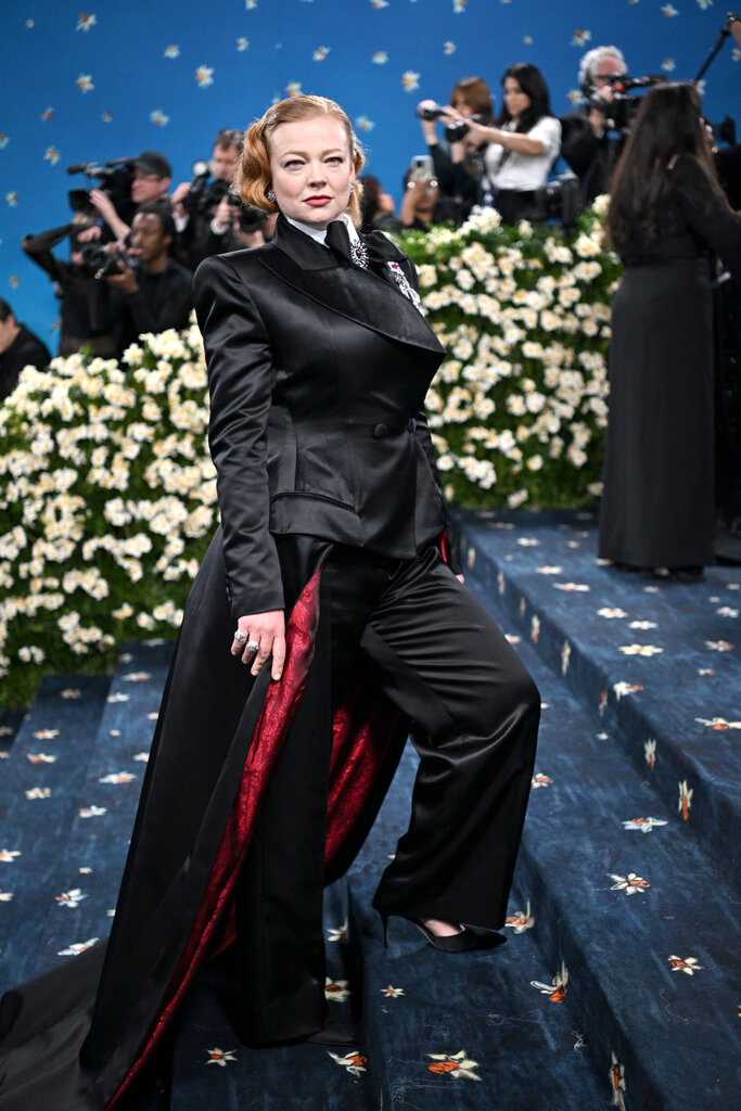 Sarah Snook wearing a black double-breasted jacket, overtop coordinating black pants and a train with red detail on the underside. 