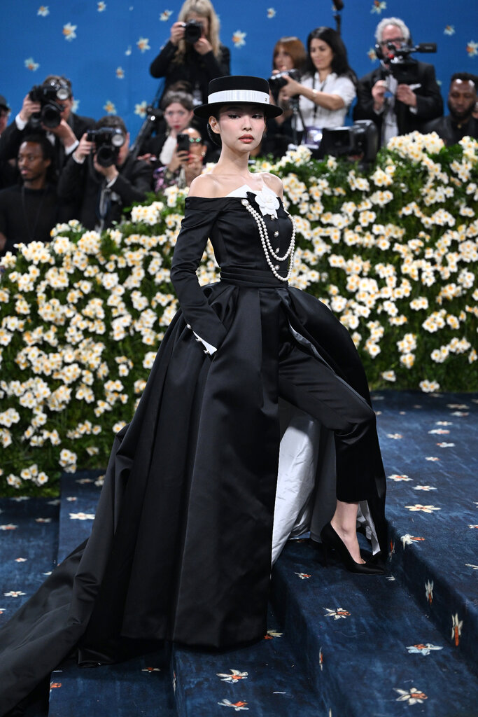 Jennie wearing a black strapless tuxedo gown with a white rosette detail at the chest, pearls and a black and white brimmed hat. 