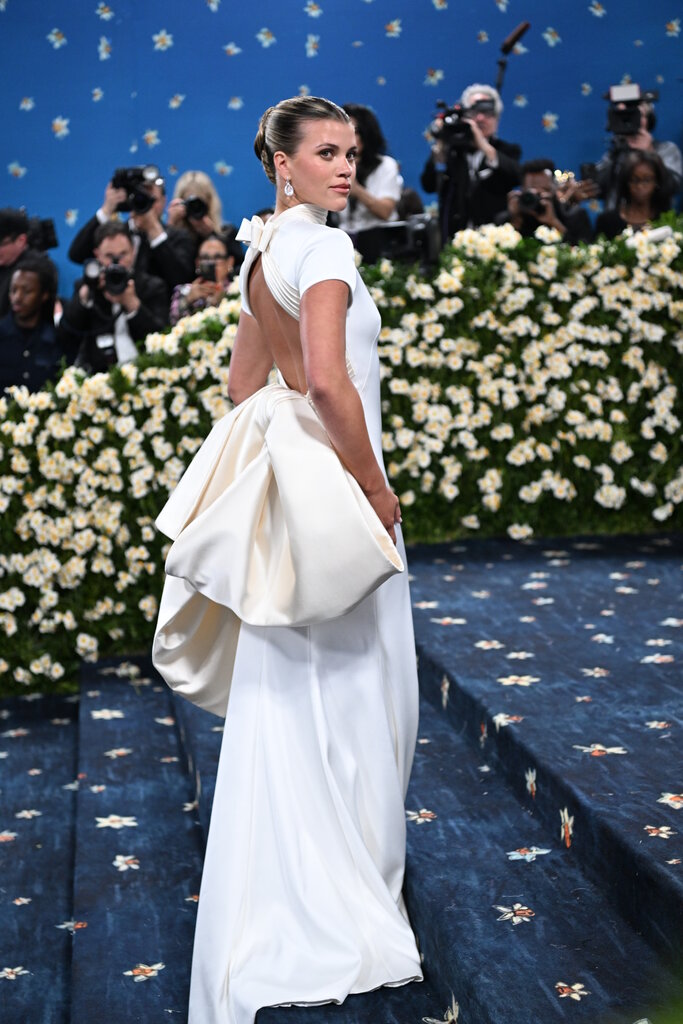 Sofia Richie wearing a long white dress with an open, upper back and cream bow on the lower back.