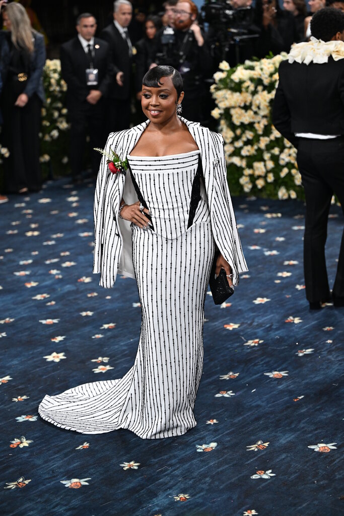 Quinta Brunson is wearing a white gown with black beaded stripes with a matching blazer that has a red rose on the lapel.