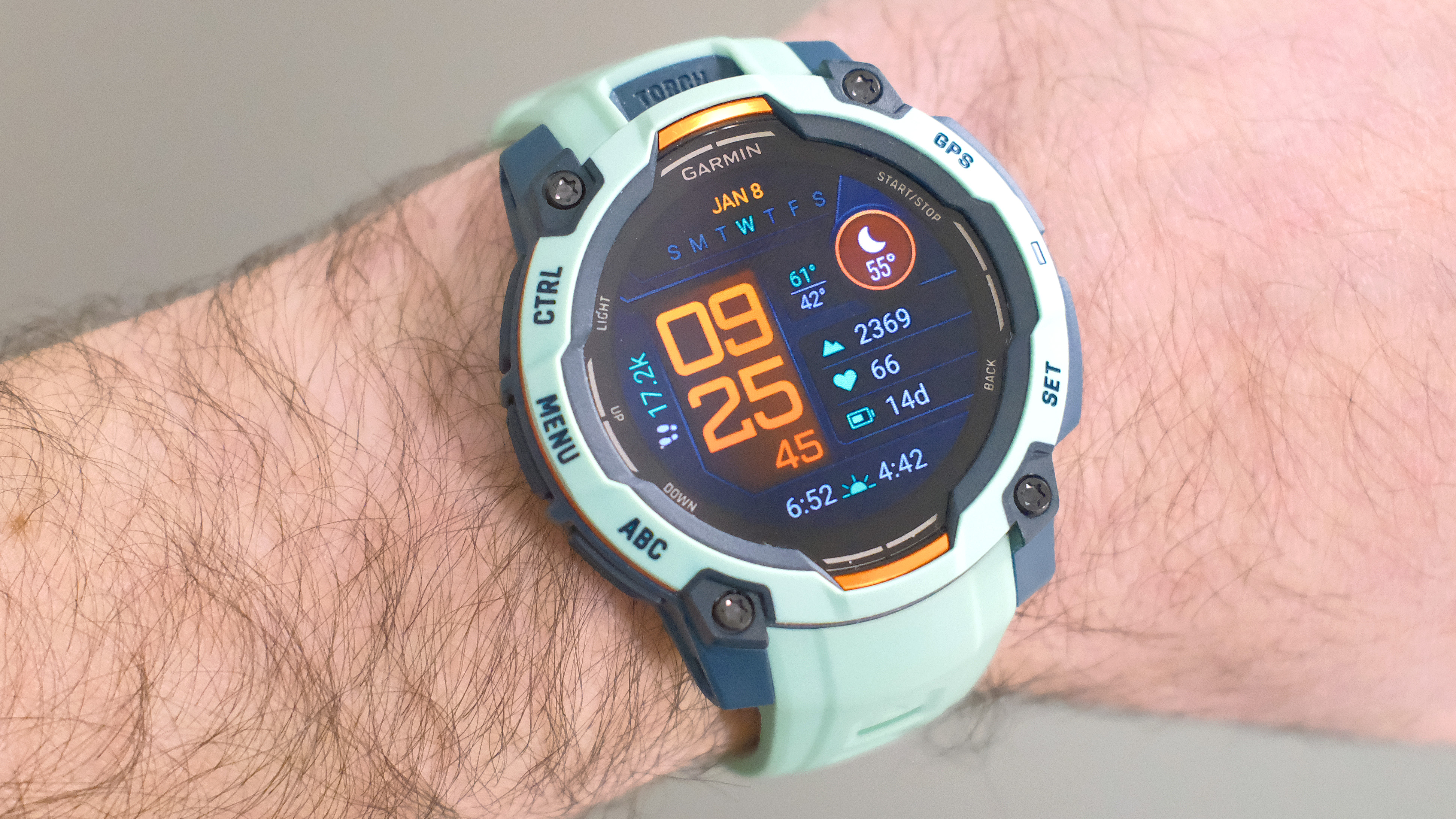 Close-up of the Garmin Instinct 3 smartwatch in orange and teal on a user's wrist with the default screen shown
