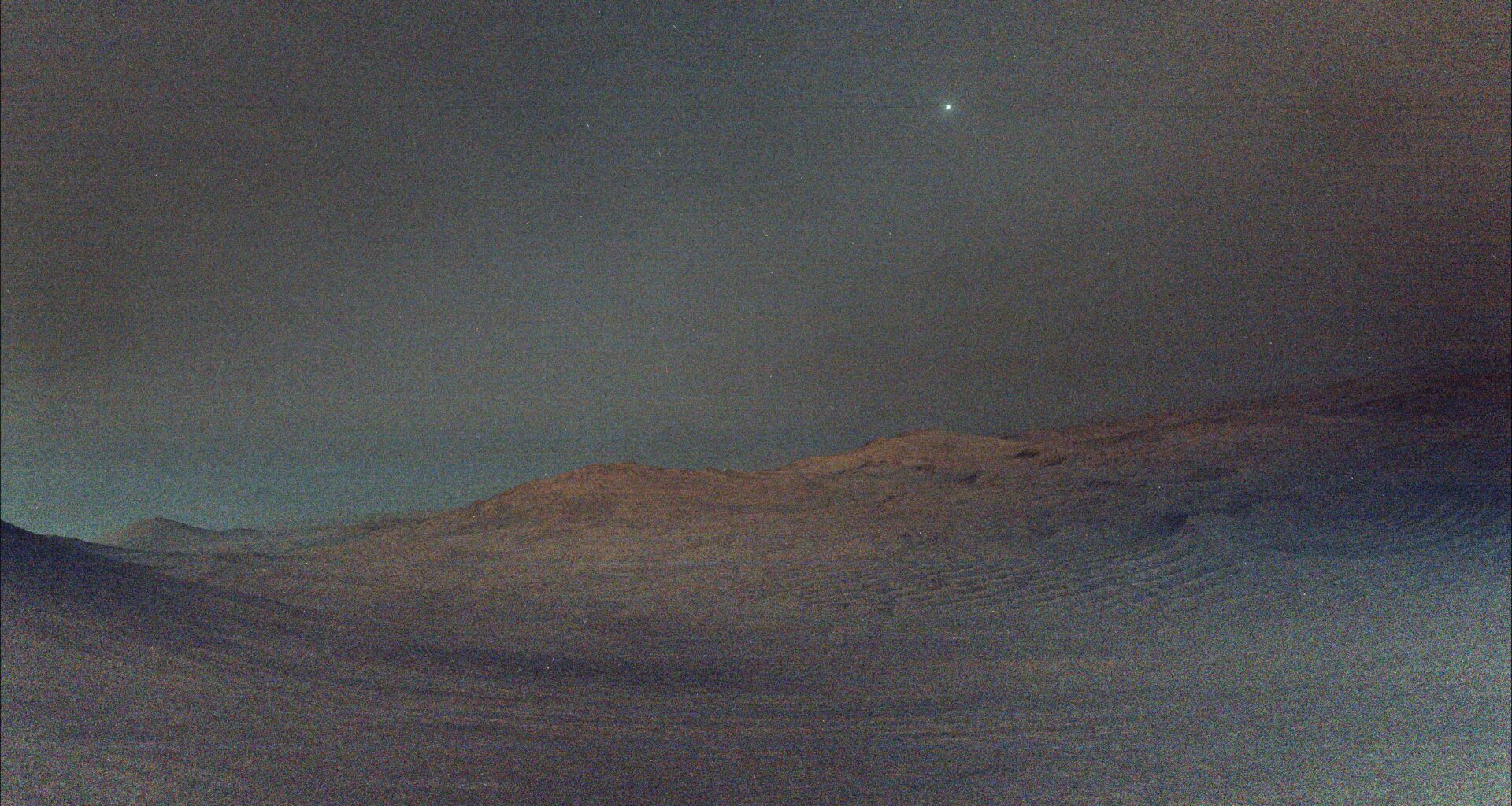 NASA's Perseverance rover captured this view of Deimos (the brightest spot in the sky), the smaller of Mars two moons, shining in the sky at 4:27 a.m. local time on March 1, 2025. The low light makes the view hazy, and the sky and ground both have a gray tint. In the distance, the crest of a crater shows Mars' signature rusty red color.