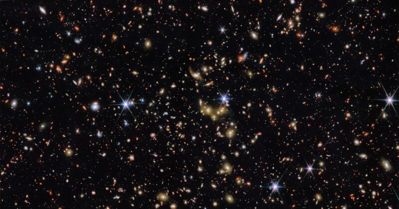 A deep space image showing countless distant galaxies, stars, and bright points of light scattered across a black background, with some stars displaying diffraction spikes.
