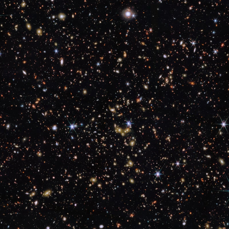 A deep space image shows numerous galaxies and stars scattered across a dark background, with bright spots, glowing clusters, and some light diffraction creating starburst effects.