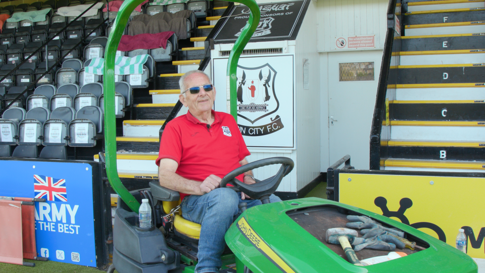 82-year-old groundskeeper Cecil Jack sitting on a John Deere riding lawnmower at Elgin City F.C. stadium.