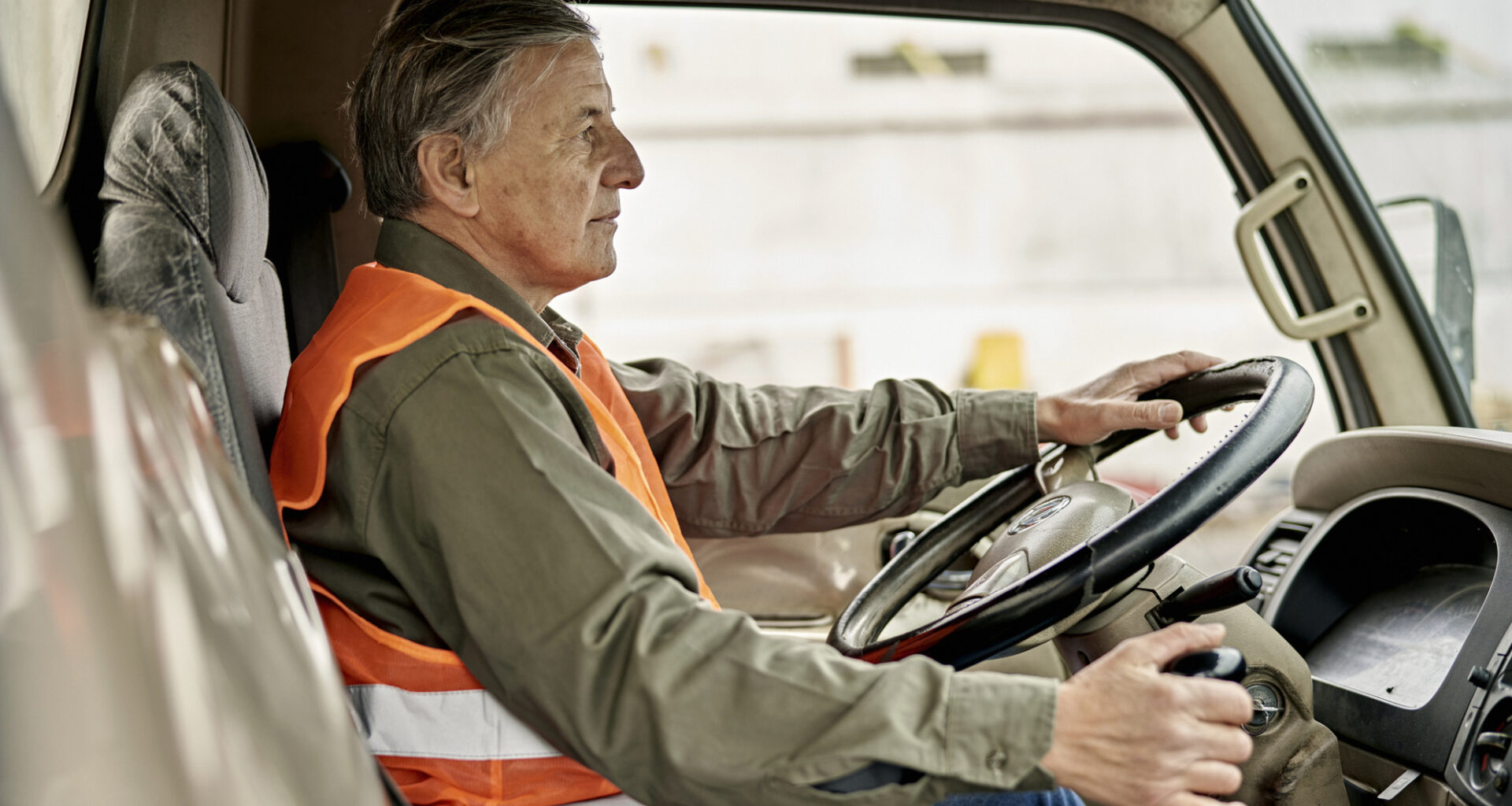 Side view of Caucasian man wearing casual clothing and reflective vest, looking straight ahead with one hand on steering wheel and other on gearshift.