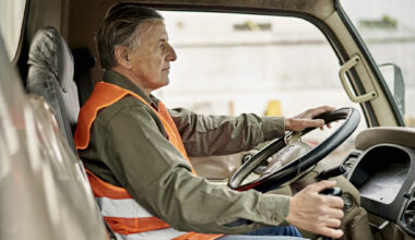 Side view of Caucasian man wearing casual clothing and reflective vest, looking straight ahead with one hand on steering wheel and other on gearshift.