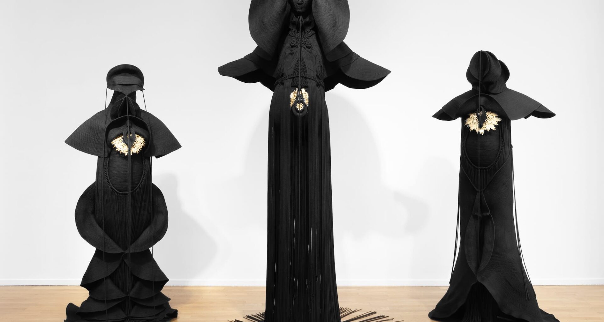 Installation view of three sculptures made from black cord, thread, and metal in the form of figures or long, dramatic garments