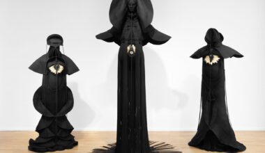 Installation view of three sculptures made from black cord, thread, and metal in the form of figures or long, dramatic garments