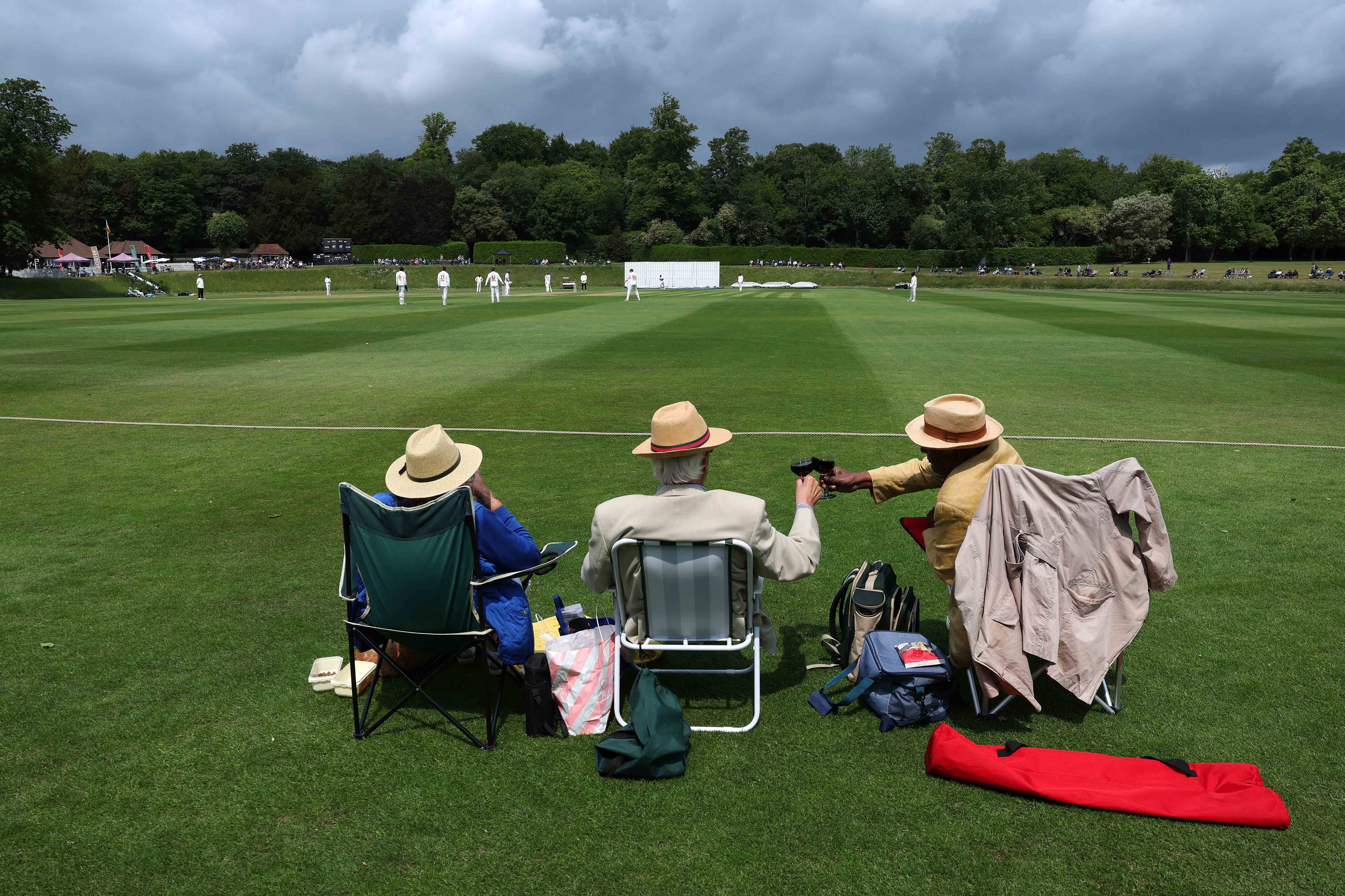 Three spectators watch a cricket match from lawn chairs.