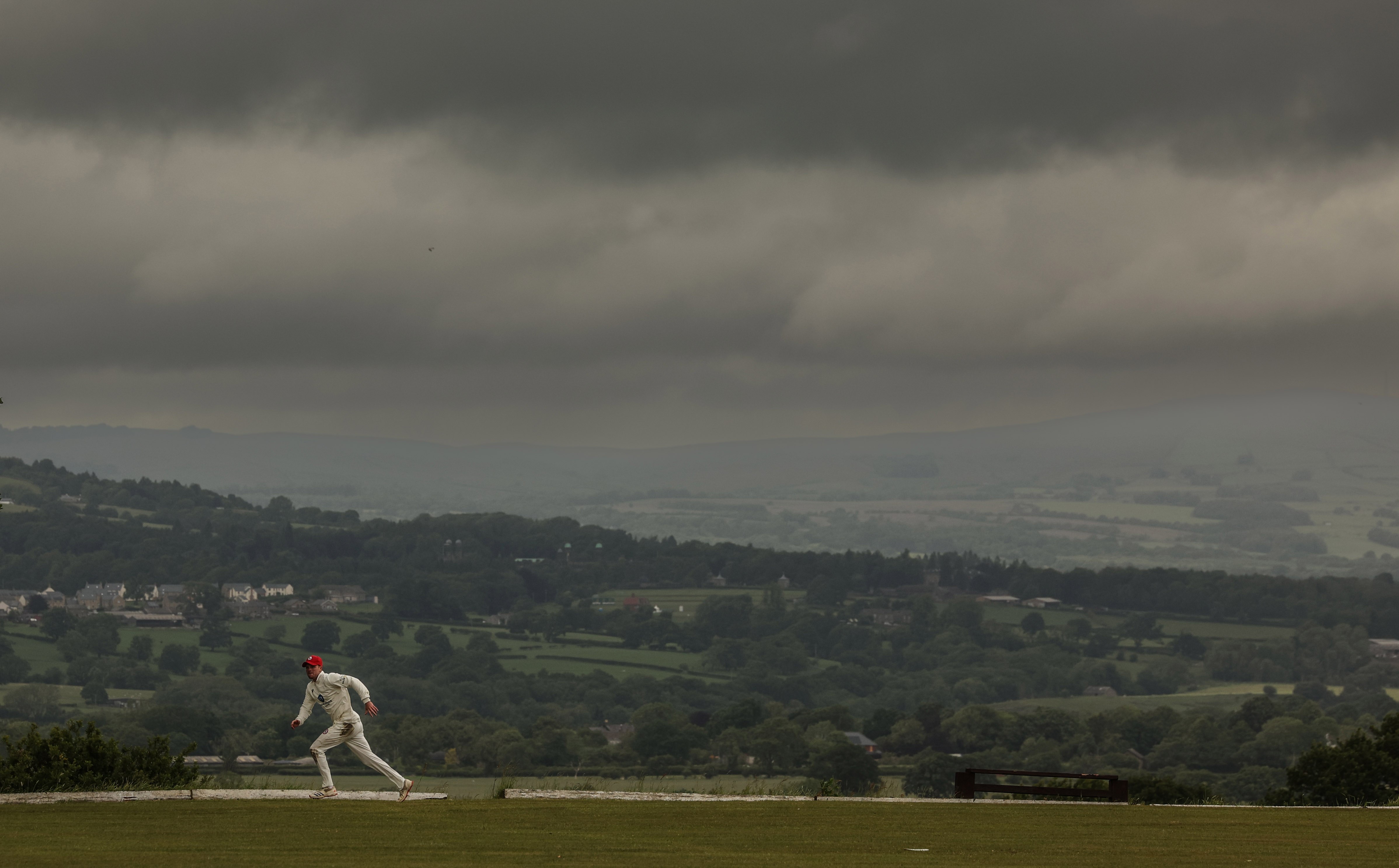 Cricket player running on a field overlooking a valley under a stormy sky.