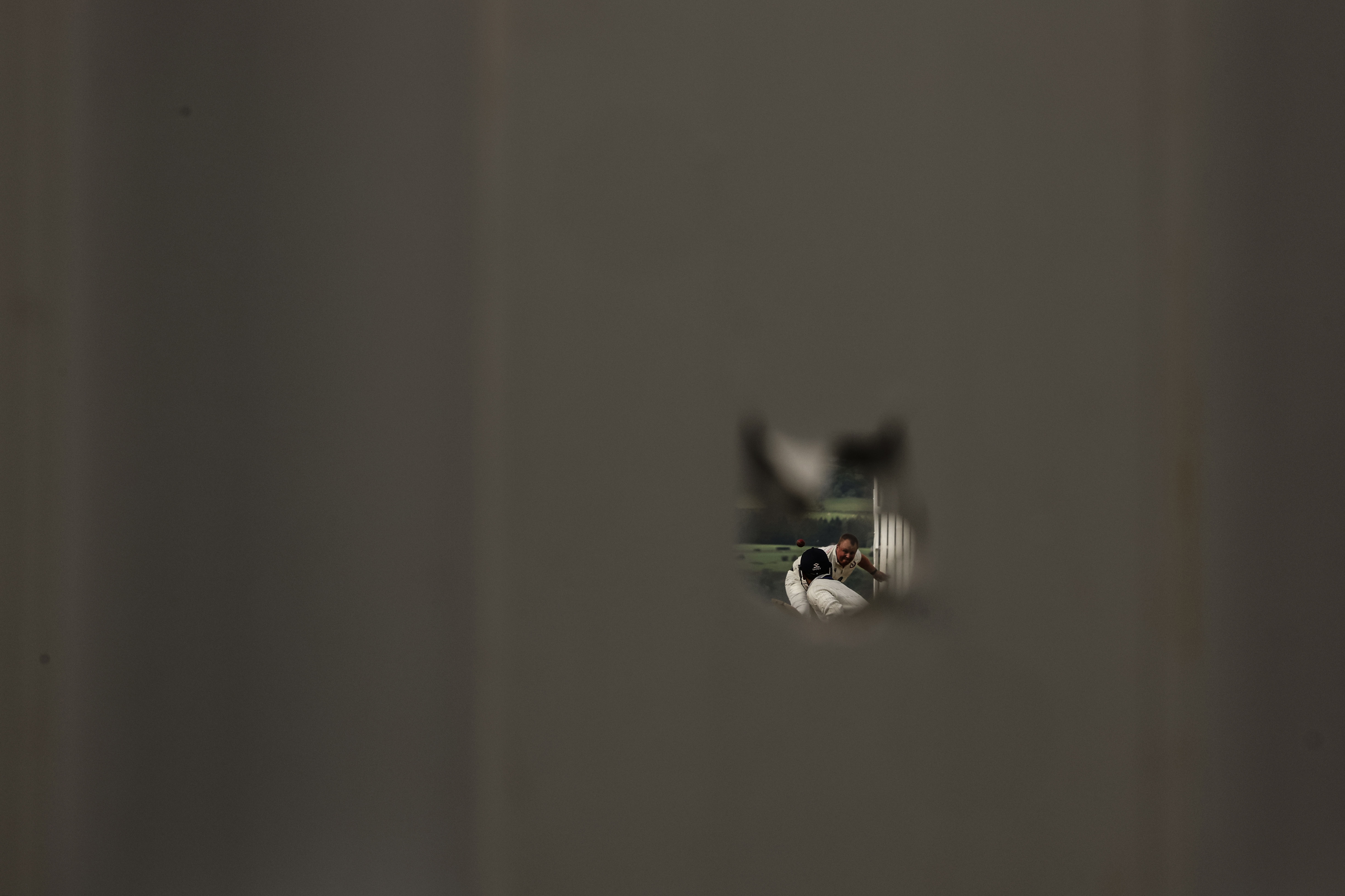Cricket match viewed through a hole in a fence.