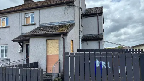 BBC A grey end-terrace house with a boarded up door and windows. There is a grey fence in front of the house and a cloudy sky above. 