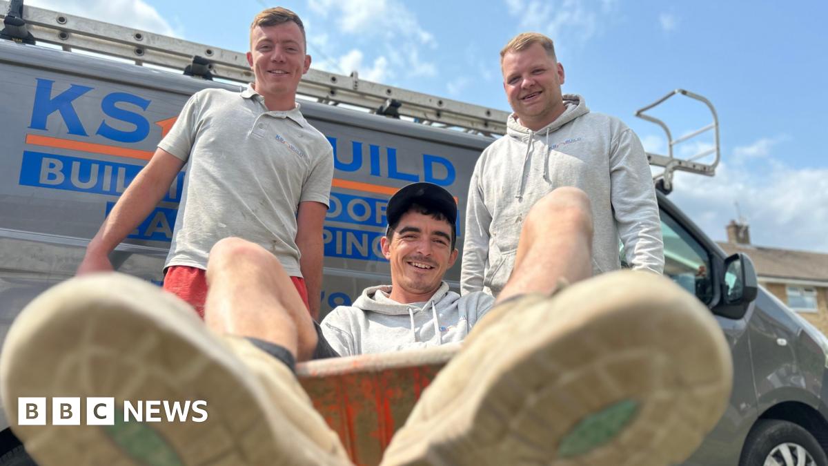 Three men, all wearing grey and smiling with one man sat in a wheelbarrow and the other two holding either handle