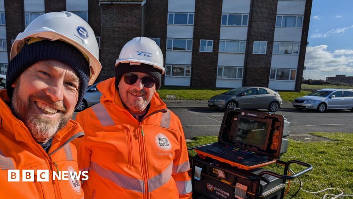 Two Southern Water team members smile for the camera next to a block of flats. They wear orange jackets and white hard hats. A high-tech laptop sits on top of equipment behind them.